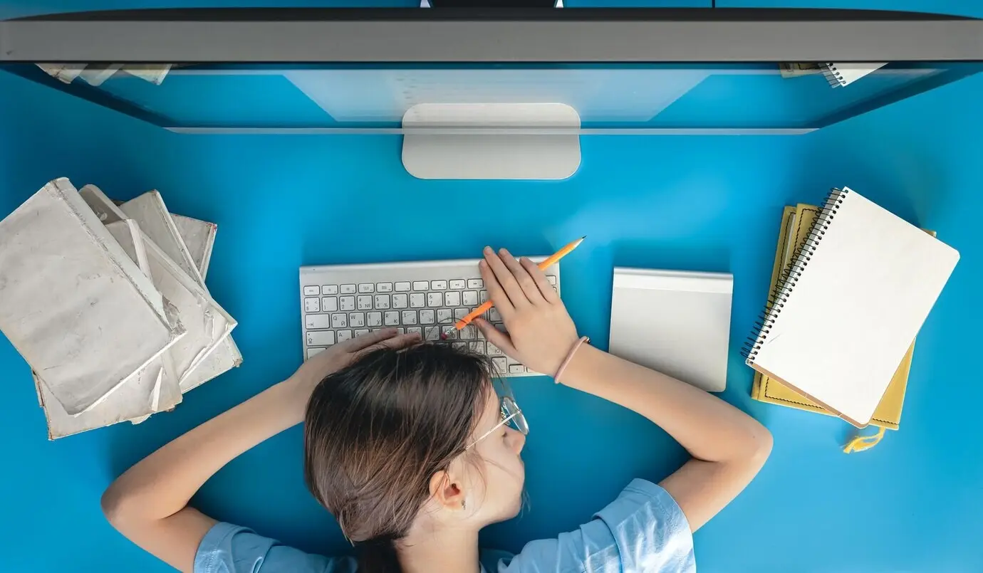 A weary girl lies on a table in front of a computer, with notepads and books.